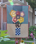 A person standing in a flower garden holds up a handmade quilt featuring a colorful geometric flower design on a blue and beige background.