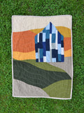 A rectangular quilted patchwork blanket featuring a stylized house and rolling hills, laid flat on green grass.