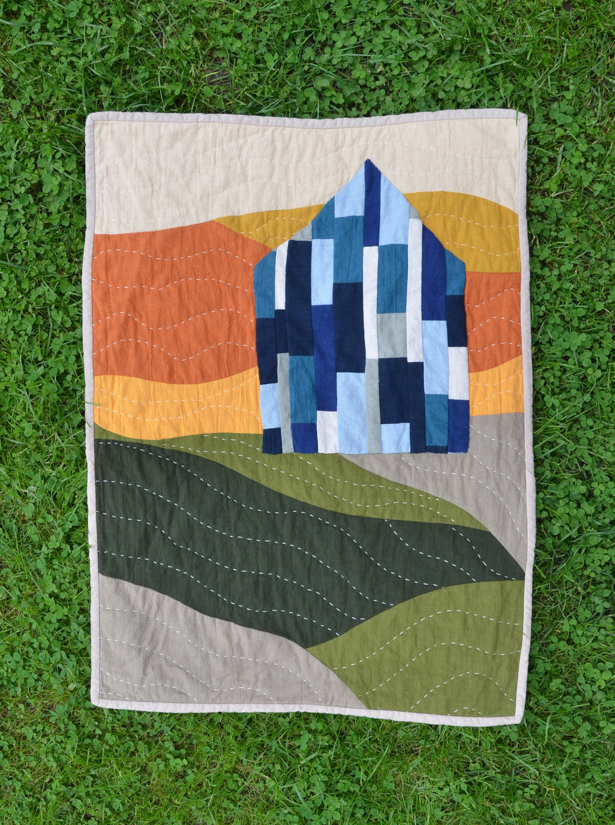 A rectangular quilted patchwork blanket featuring a stylized house and rolling hills, laid flat on green grass.